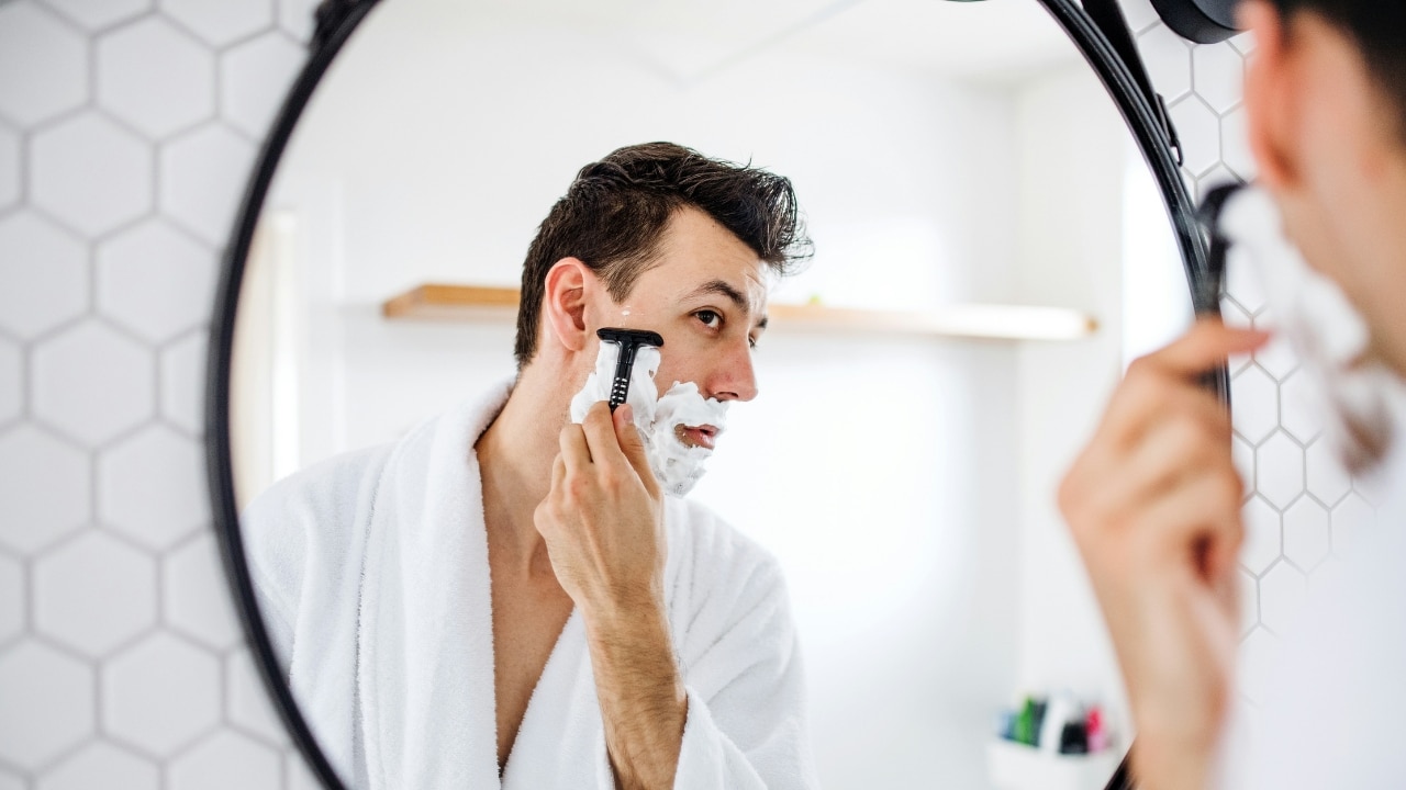 A man in a white robe shaves his face with a razor in a mirror.
