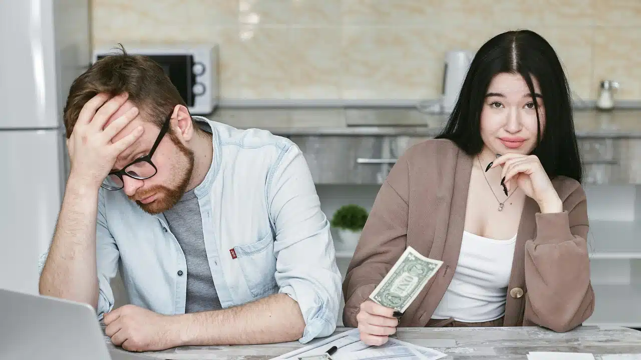 A man looks down at a laptop while a woman holds a one-dollar bill nearby.