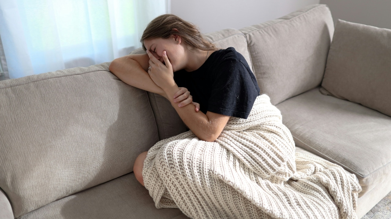 A woman sits on a couch under a blanket, covering her face with her hand.