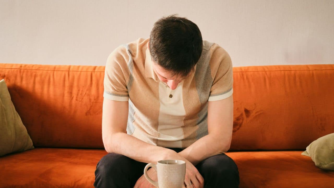 A man sits on an orange sofa with his head bowed over a grey mug.