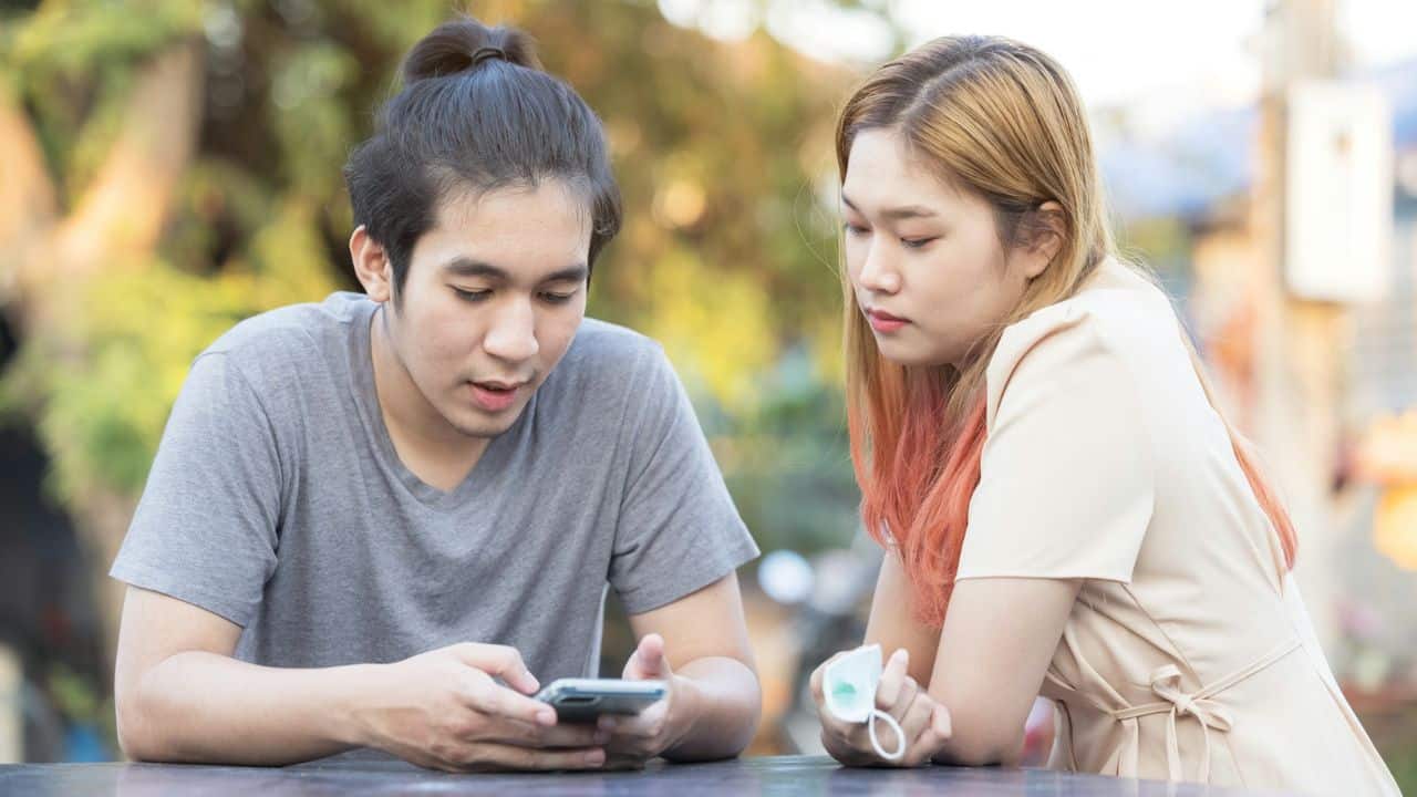 A man looks at a smartphone while a woman sits beside him looking downward.