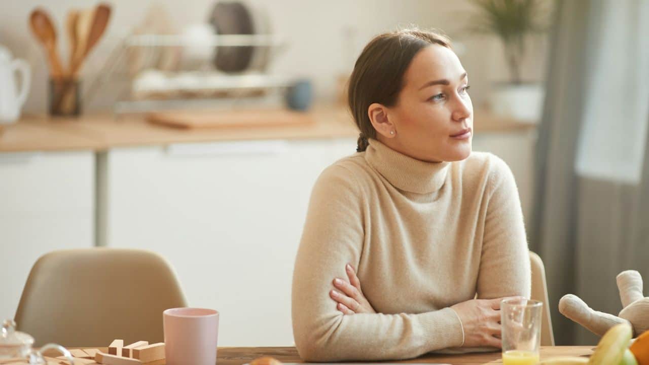 A woman in a beige turtleneck sits at a table and looks off to the side.