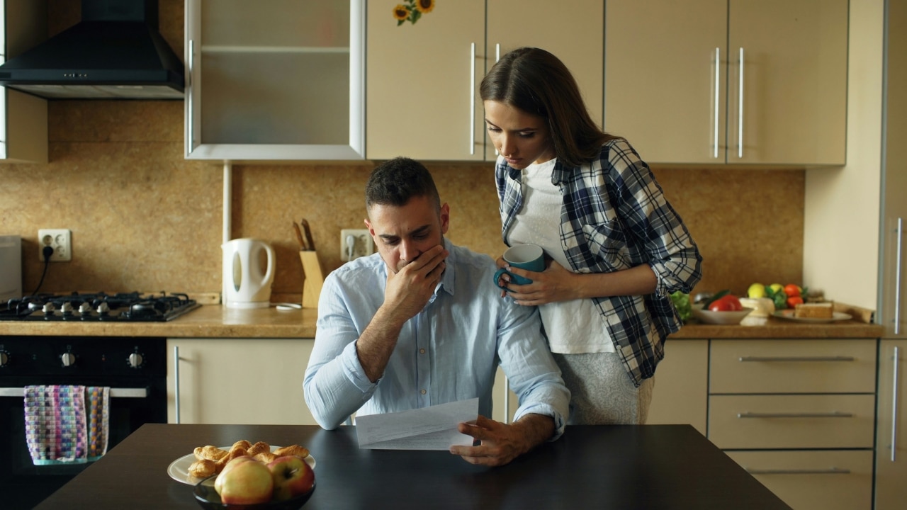 A woman holding a mug stands over a seated man reading a document in a kitchen.