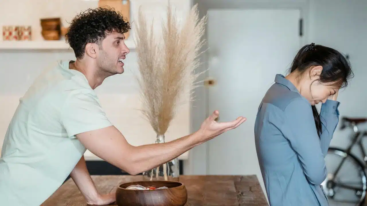 A man gestures with an open hand toward a woman who is covering her face.