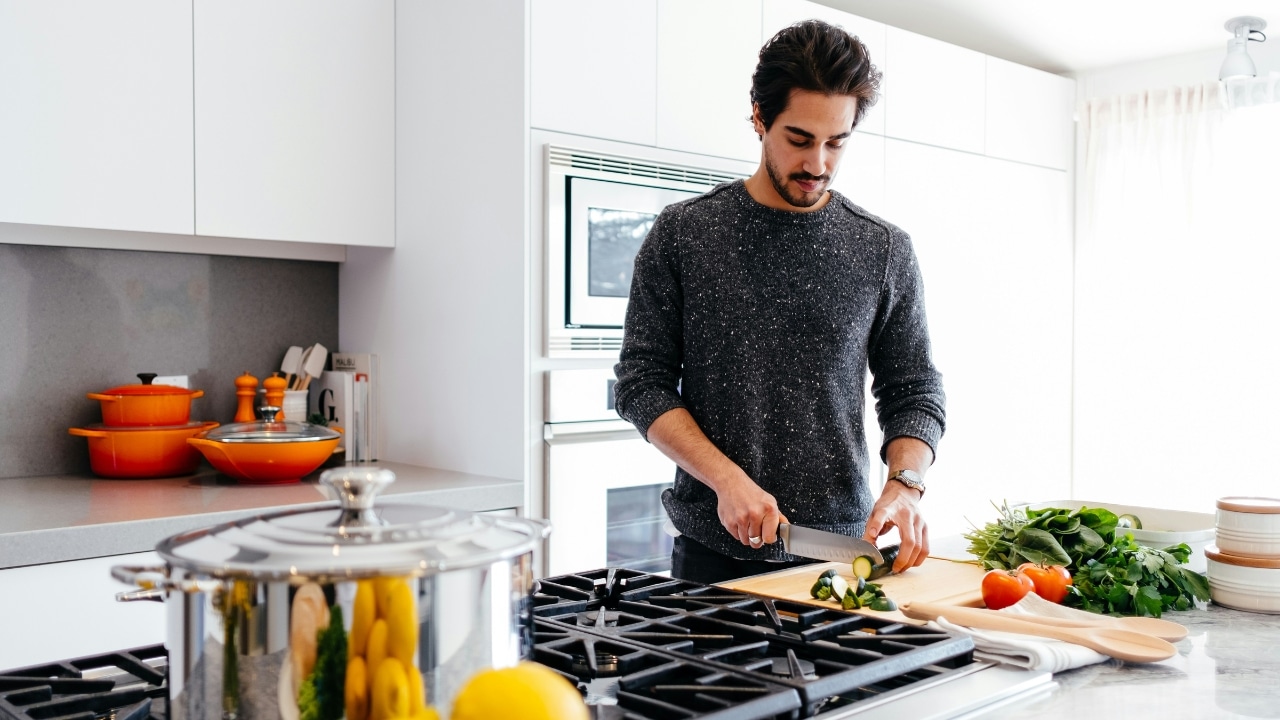 A man in a grey sweater slices a cucumber on a wooden cutting board in a kitchen.