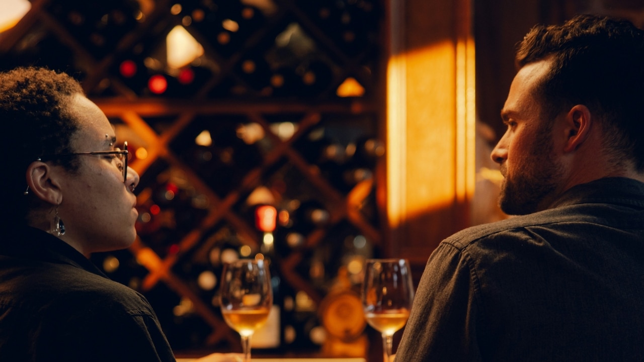 Two people sit at a bar with glasses of wine in front of a wine rack.