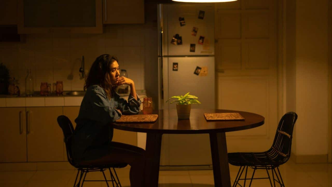 A woman sits alone at a round wooden table in a dimly lit kitchen at night.