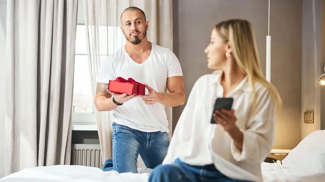 A man kneels holding a red gift box while a woman looks at her phone.
