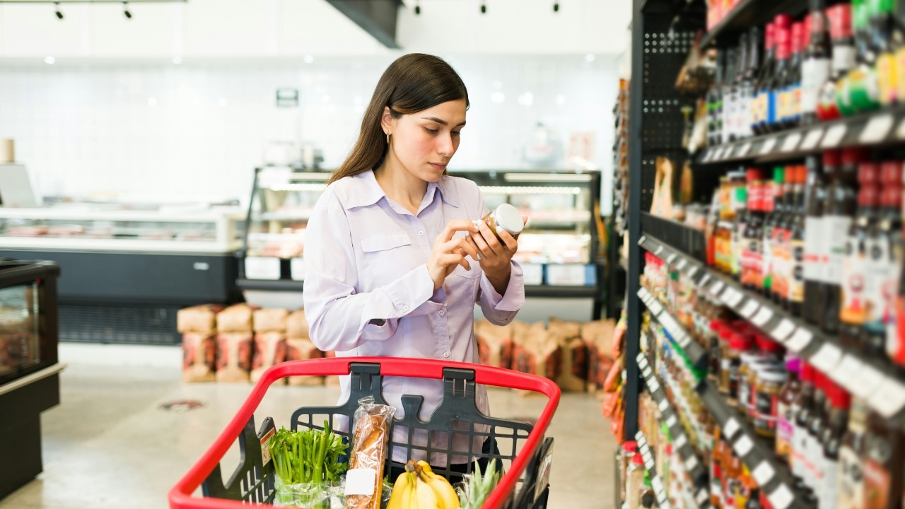 A woman in a purple shirt looks at a jar while standing with a grocery cart.