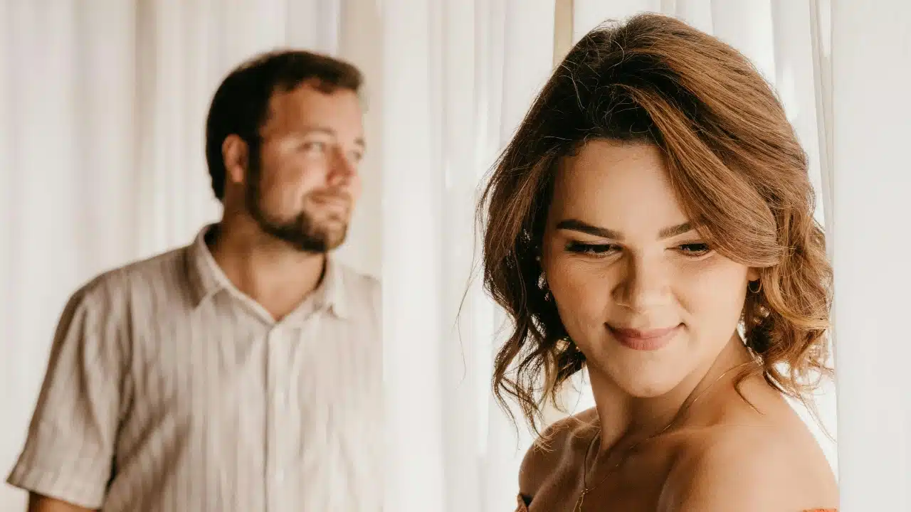 A woman smiles in the foreground while a man stands slightly blurred behind her near white curtains.