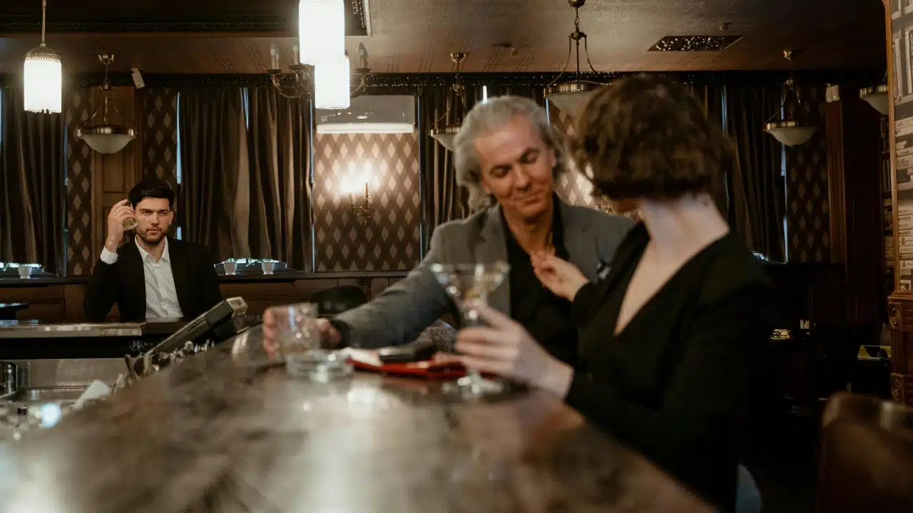A man in a suit watches a couple sitting and talking at a dimly lit bar.