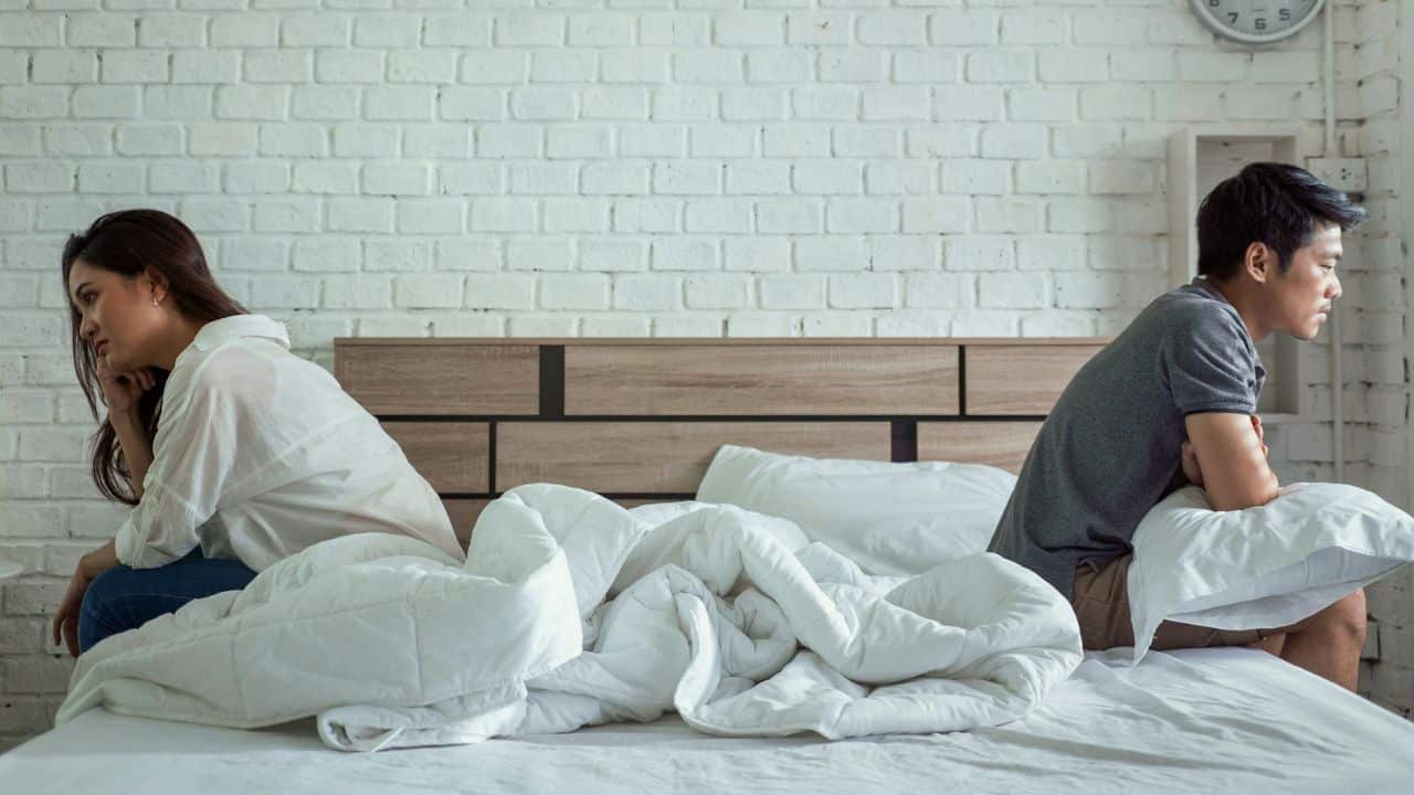 A man and woman sit on opposite sides of a bed with their backs to each other.