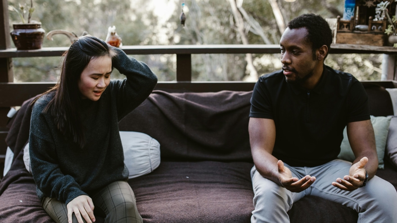 A man speaks with open palms while a woman beside him touches her head.