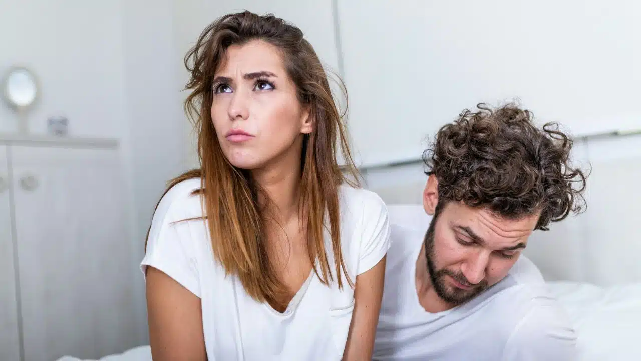 A woman with a frustrated expression looks upward beside a man looking down on a bed.