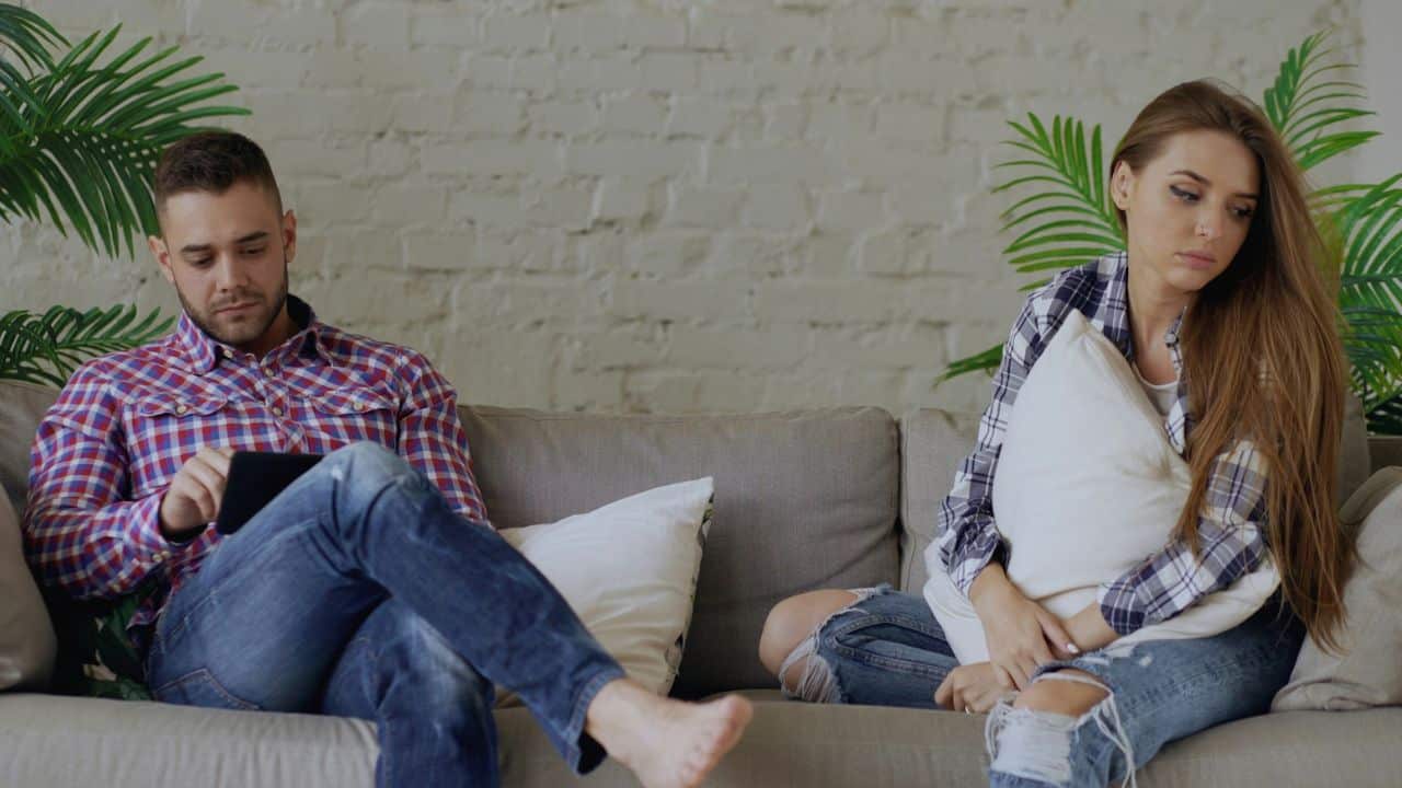A man uses a tablet while a woman sits beside him clutching a white pillow.