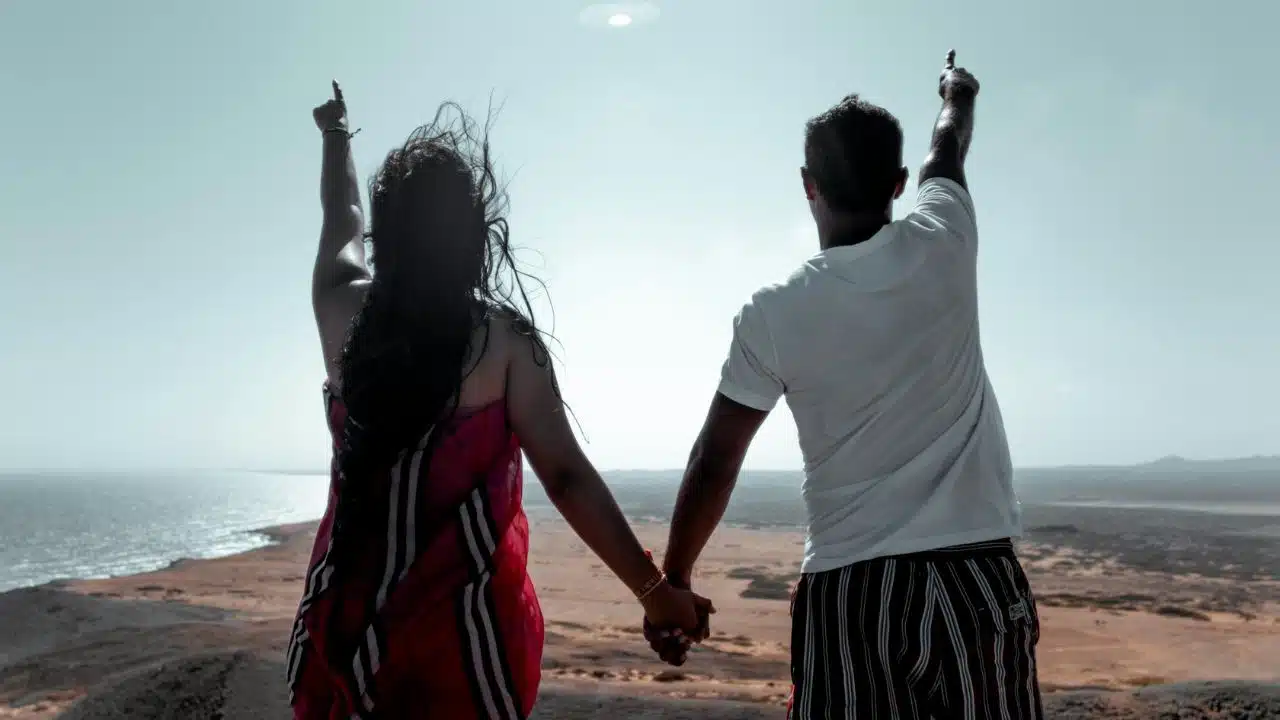 A man and woman stand back-to-camera, holding hands and pointing toward the sky.