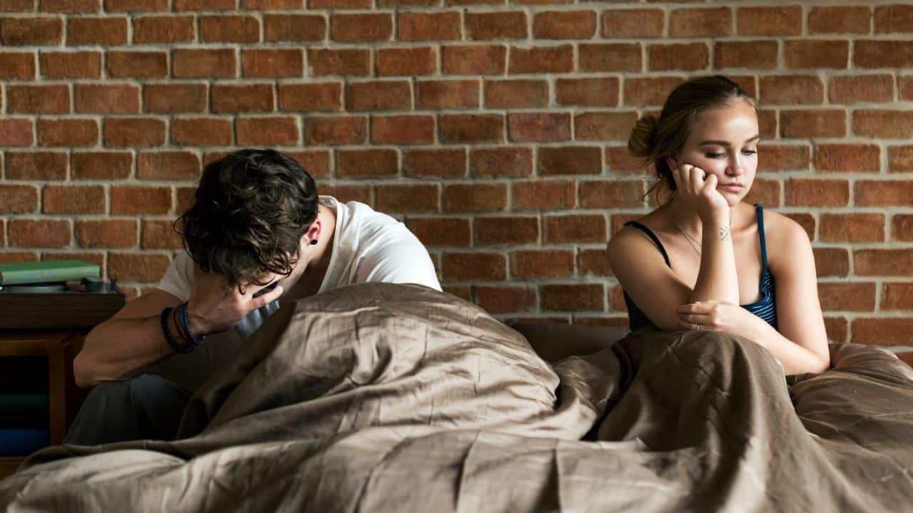 A man and woman sit in a bed against a brick wall looking in opposite directions