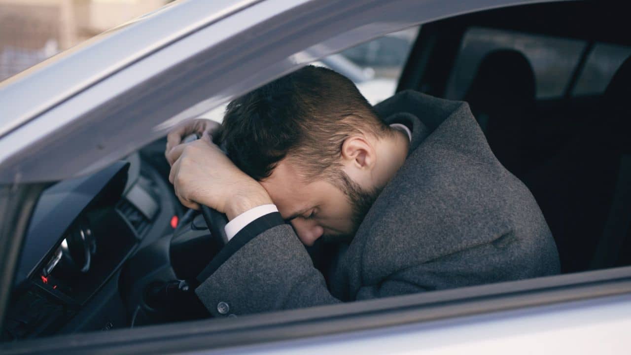 A man in a grey coat rests his head on a car steering wheel inside.