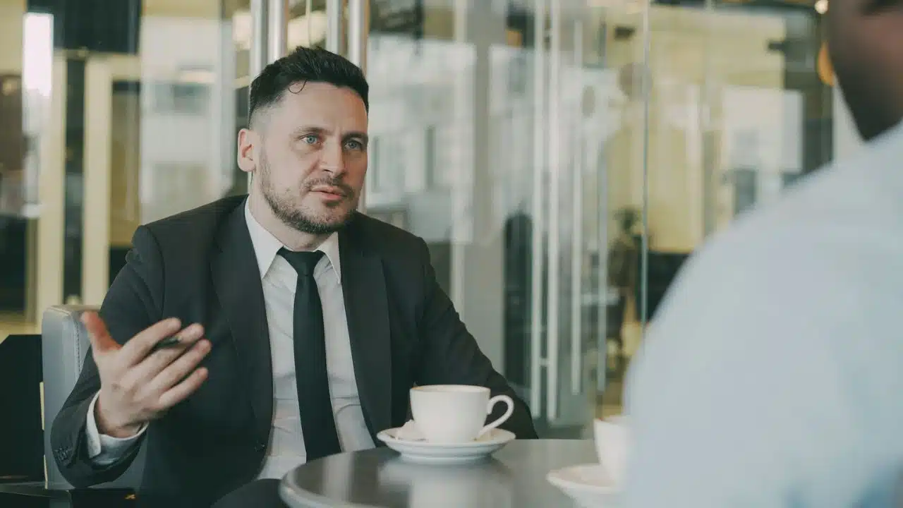 A man in a suit gestures while talking to someone across a table with coffee cups.