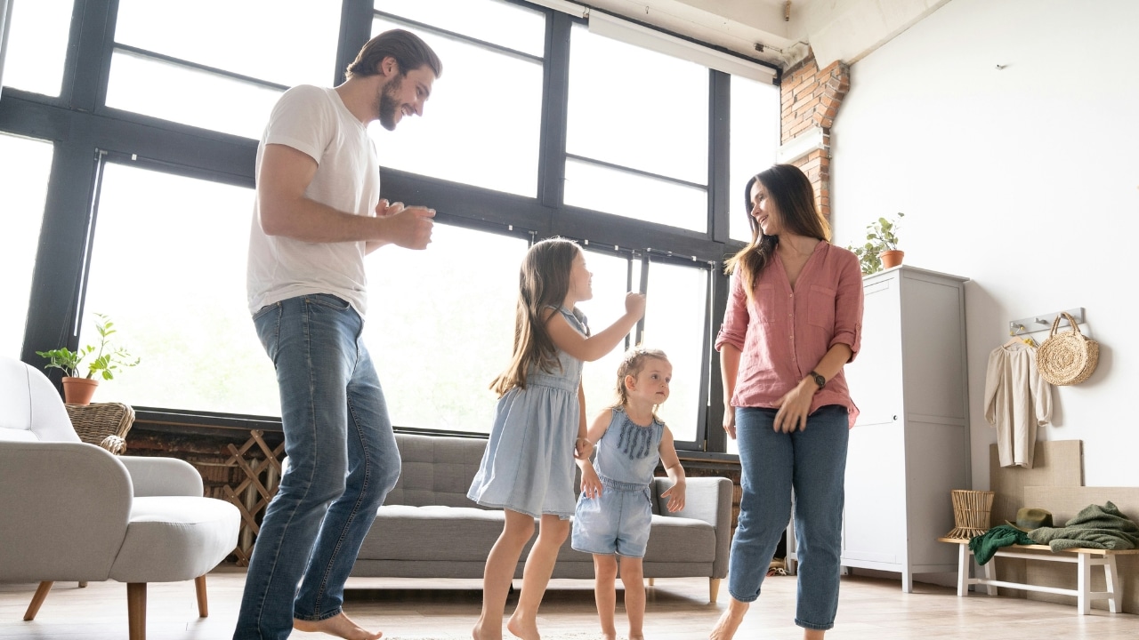 A man, woman, and two young children dance together in a bright, sunlit living room.