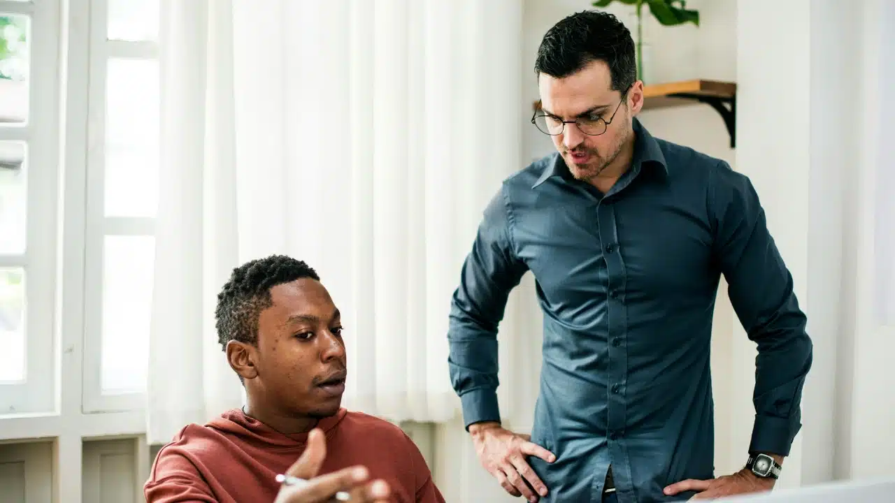 One man stands with hands on hips while another man sits and speaks to him.