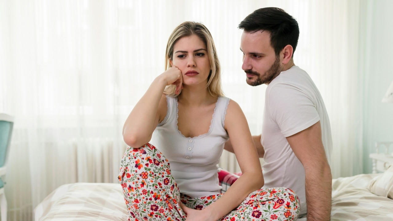 A woman in floral pants sits on a bed looking away as a man watches her.