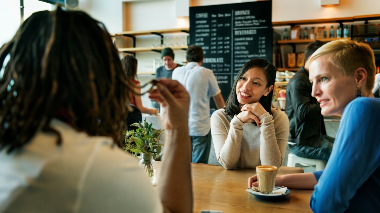 Two women sitting at a cafe table talking, with the back of another person's head visible.