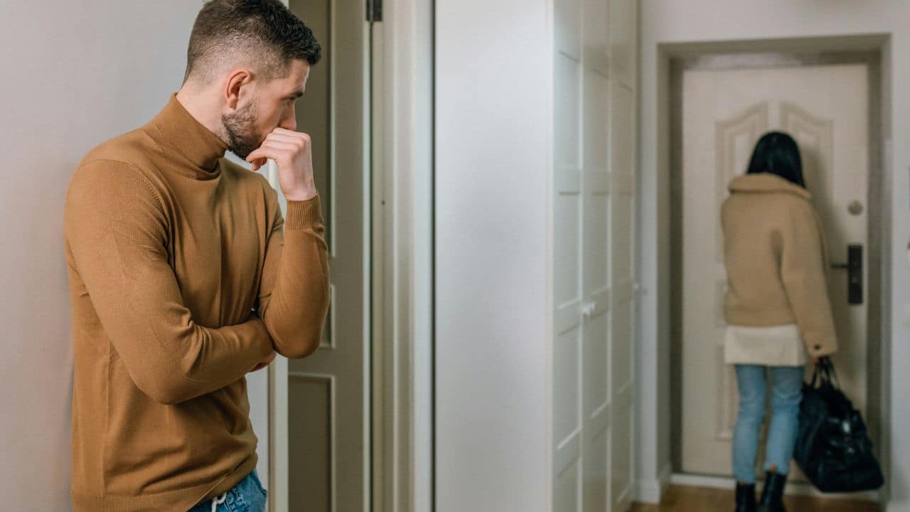 A man watches from a hallway as a person with a bag stands by a door.
