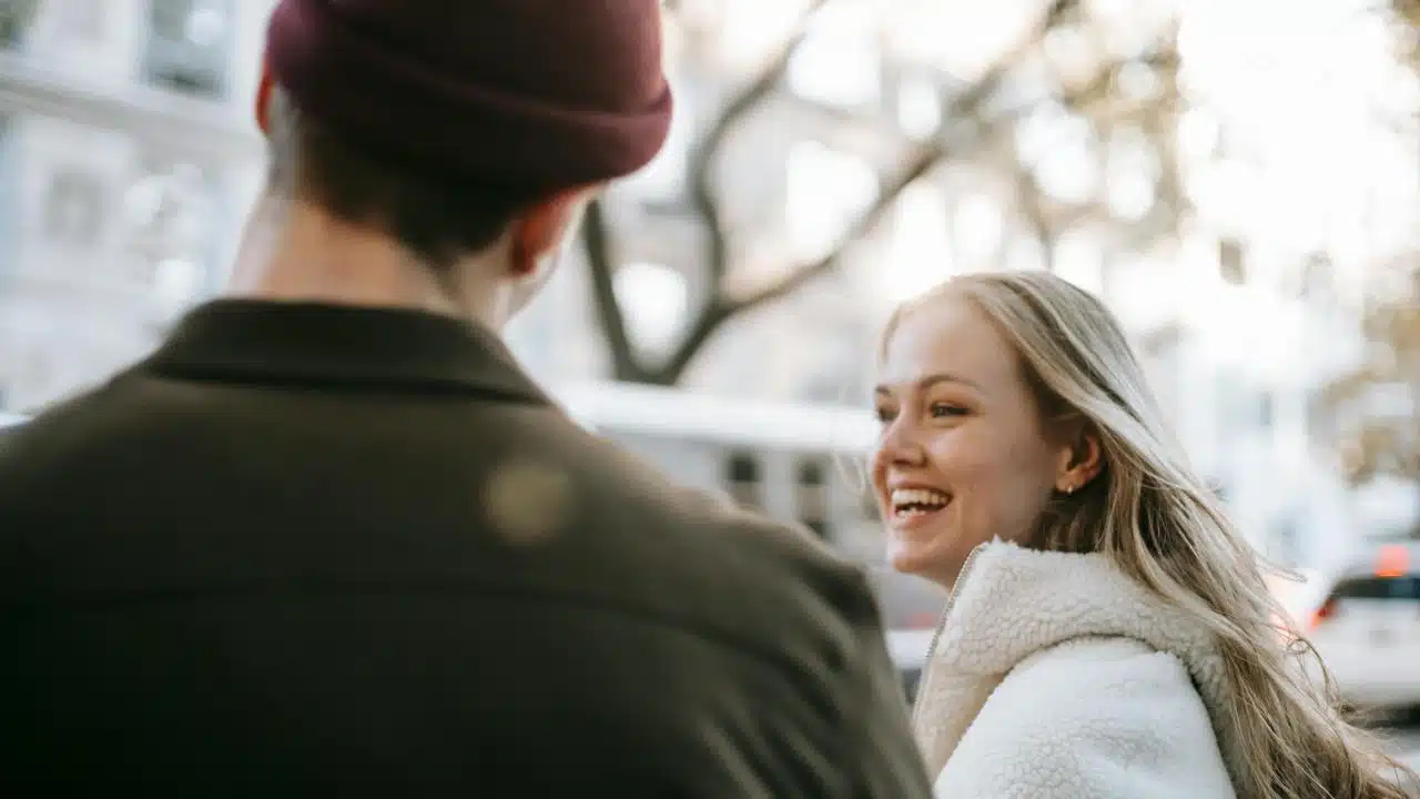 A woman smiling while talking with a person outdoors in a city setting.