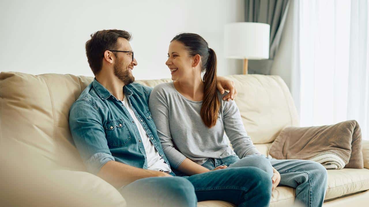 A couple sits on a couch smiling and looking into each other’s eyes.