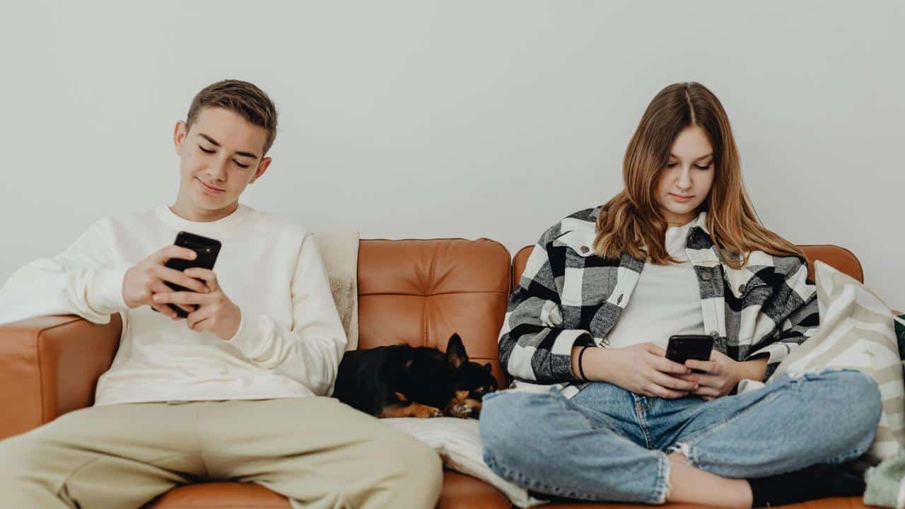 A couple sitting on a couch using their phones with a dog between them.