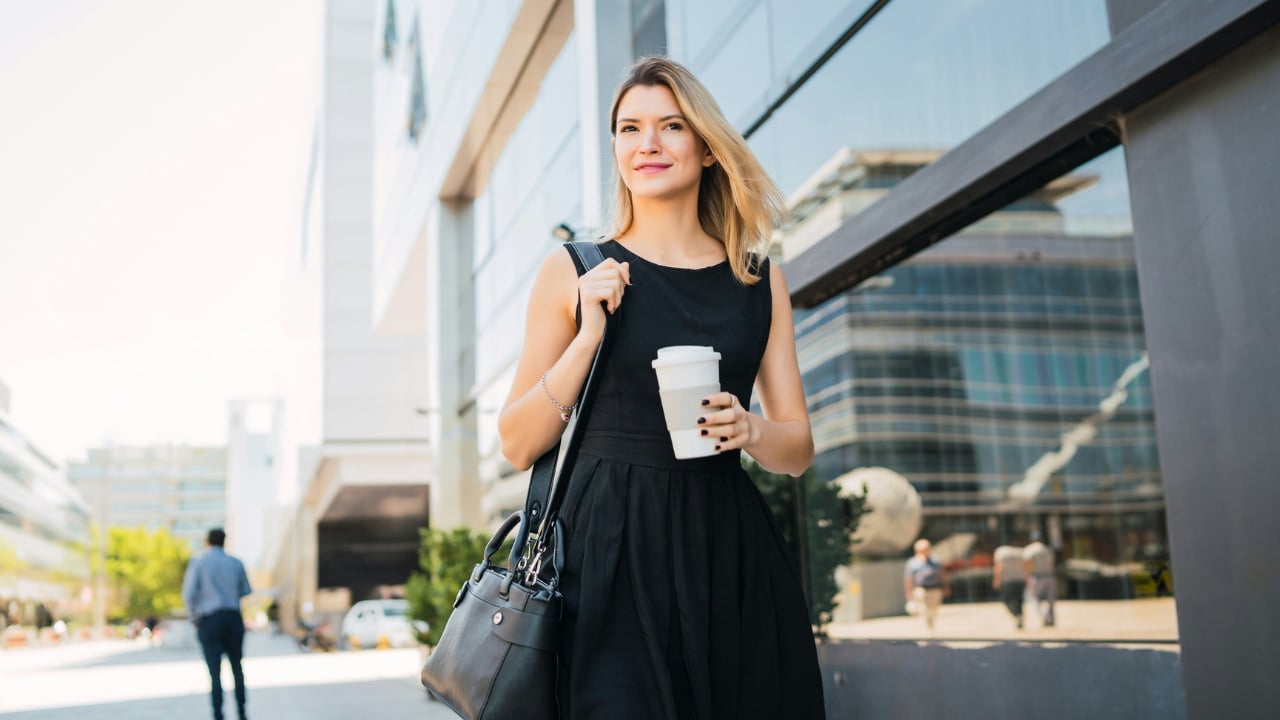 Woman in a black dress carrying a shoulder bag and a white coffee cup outdoors.