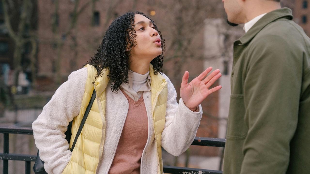 A woman with curly hair gestures with one hand while speaking closely to a man outdoors.