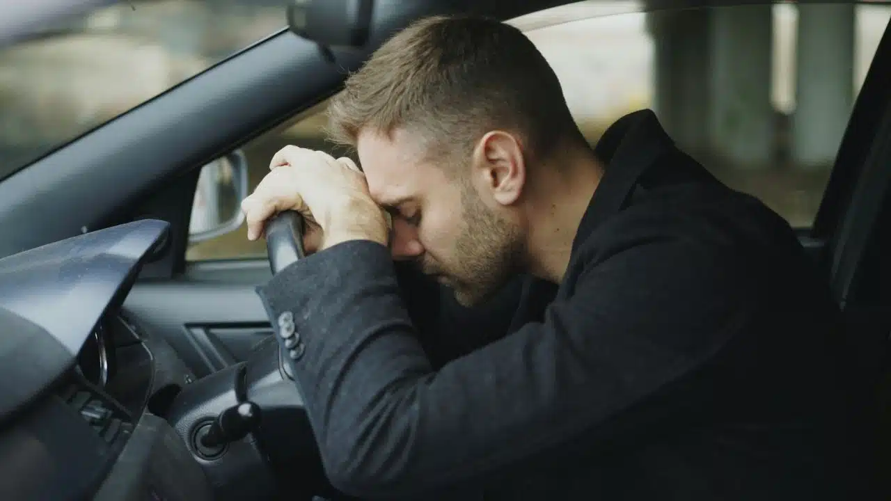 A man in a dark jacket rests his head on a car steering wheel indoors.
