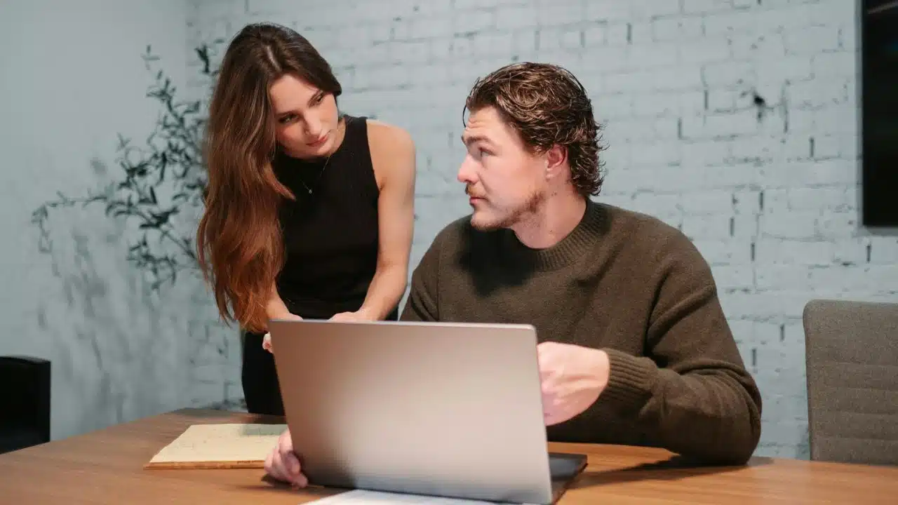 A woman leans over a man's shoulder as he works on a laptop at a desk.
