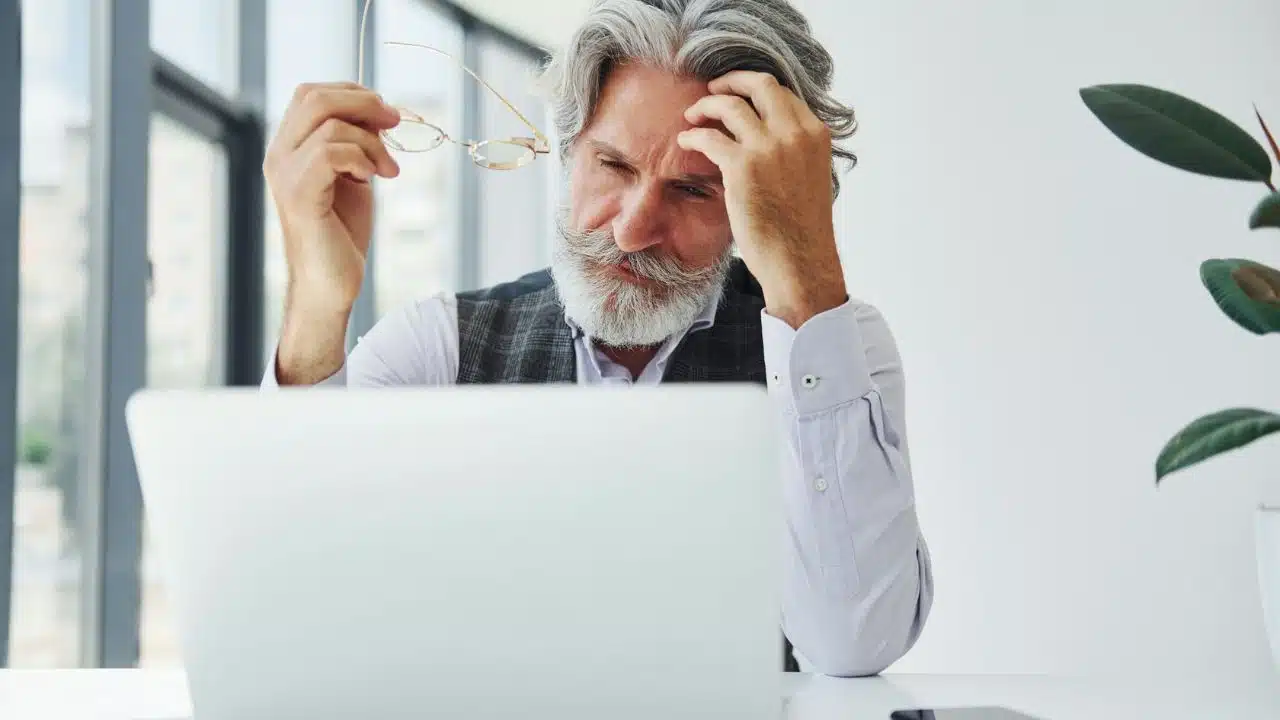 An older man with a mustache holds his glasses and touches his head while looking down.