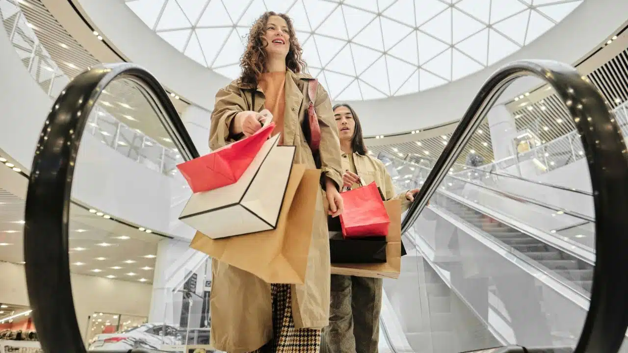 A woman and a man descend an escalator while carrying several shopping bags each.