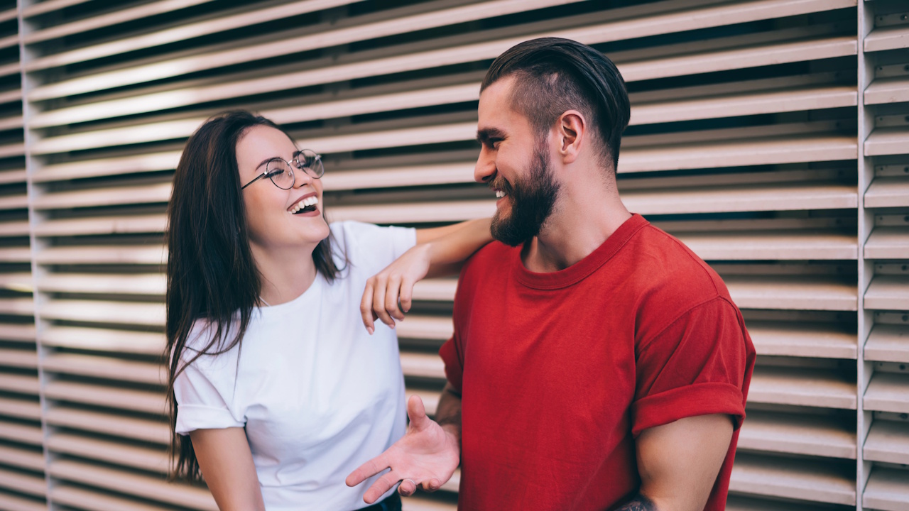 A smiling couple leaning against a slatted wall, the woman wearing glasses.