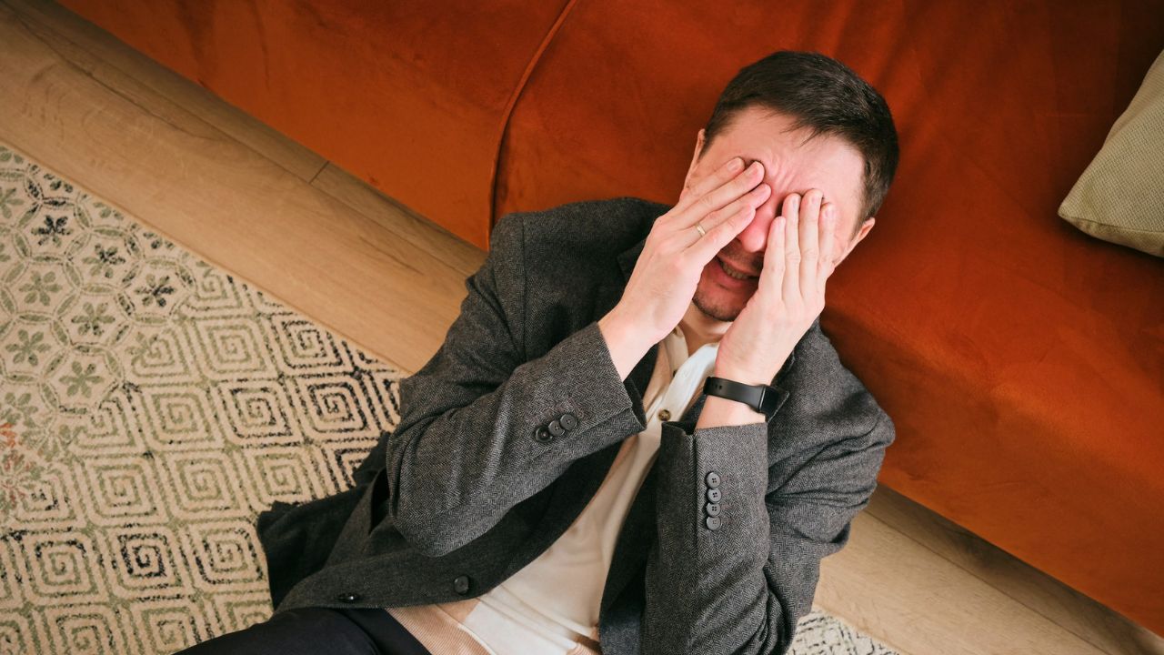 An upset man lying on the floor near a sofa, covering his face and crying.