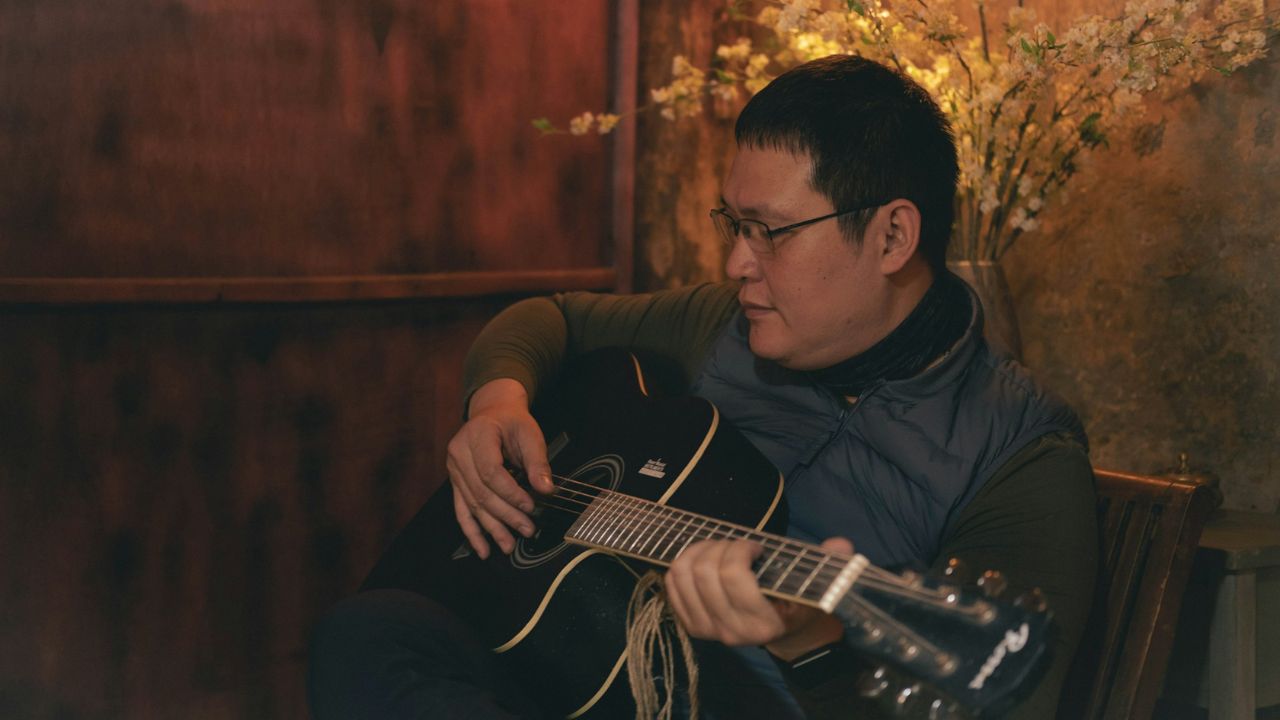A man wearing glasses and a vest is playing a black acoustic guitar indoors.