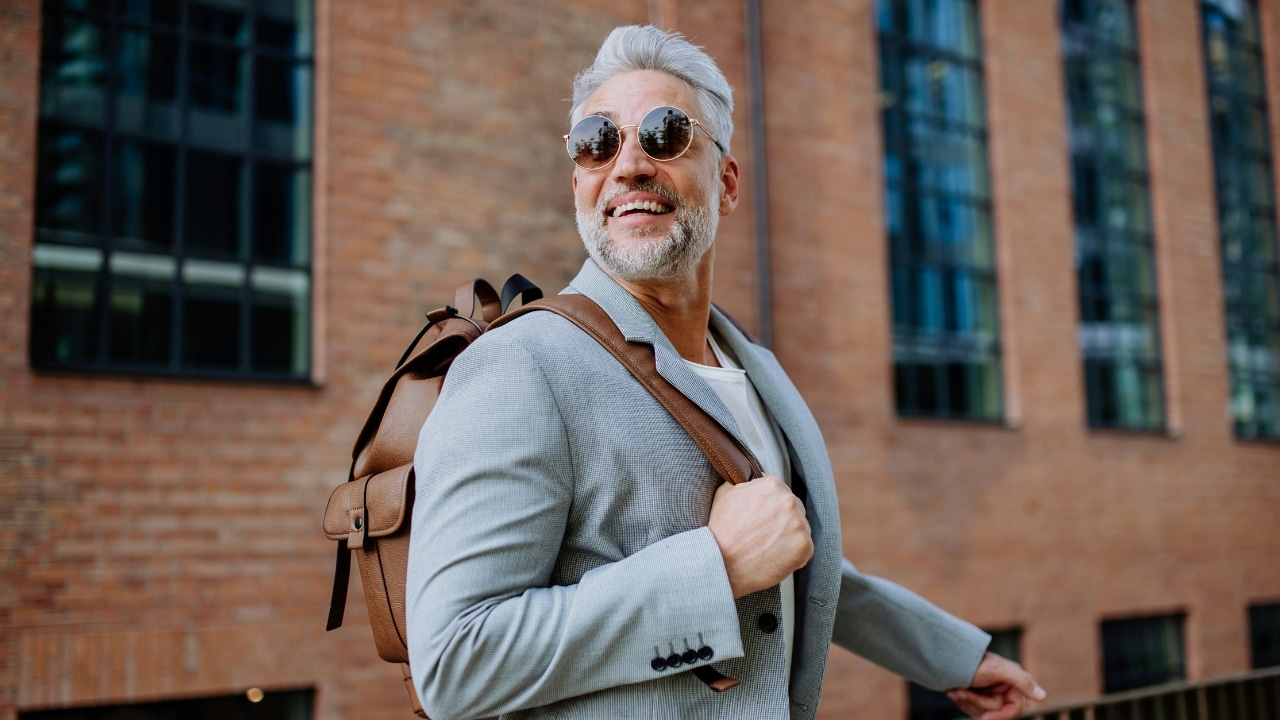 A smiling man with gray hair wears sunglasses and a backpack near a brick building.