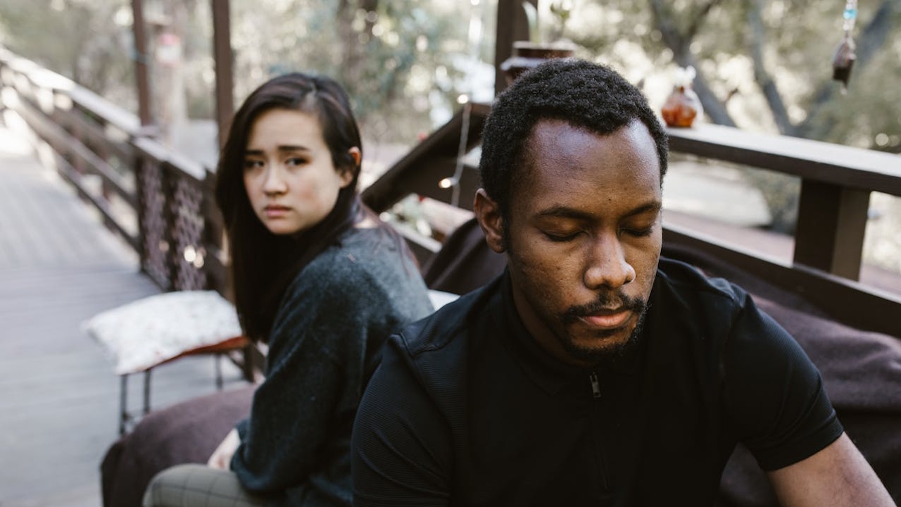 A man with a serious expression and a woman looking upset sitting separately outdoors.