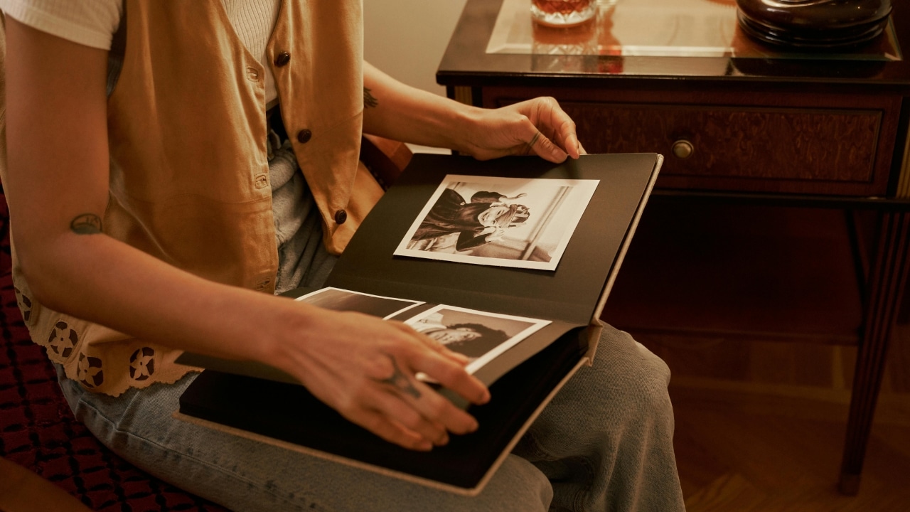 A person with hand tattoos flips through a black photo album while sitting in a chair.