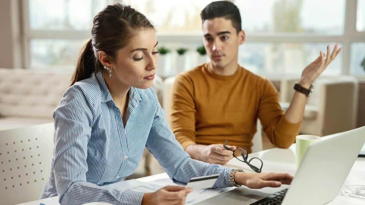 A woman holds a card while using a laptop as a man watches her.