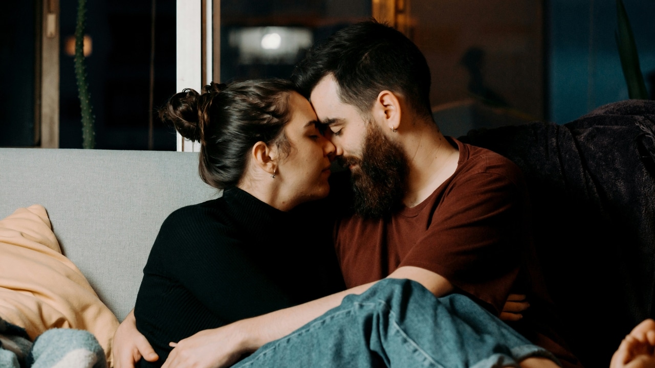 A man and woman sit closely on a sofa with their foreheads touching.