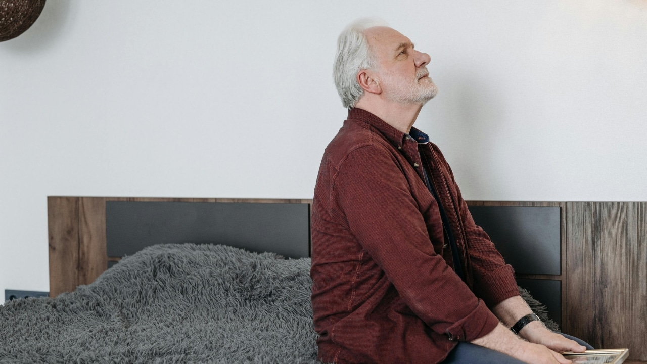 An older man in a maroon shirt sits on a bed and looks upward.