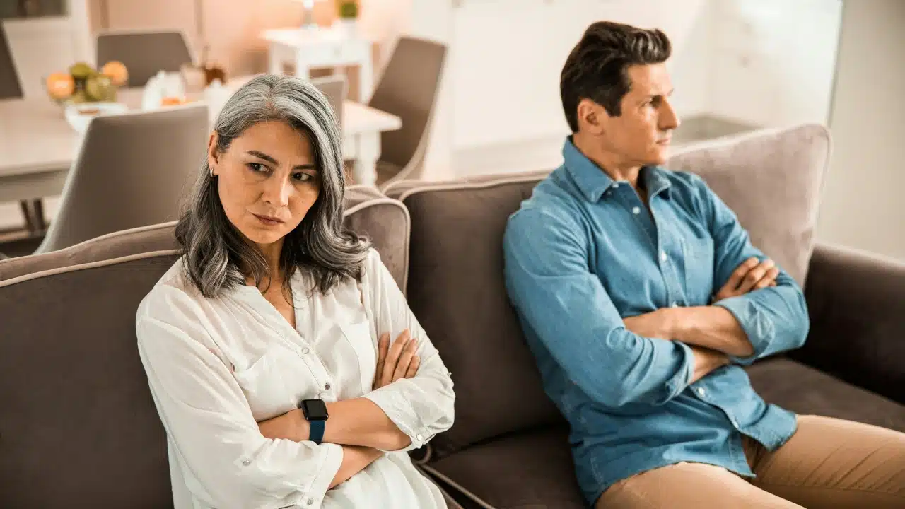 A woman and a man sit on a couch with their arms crossed looking away.