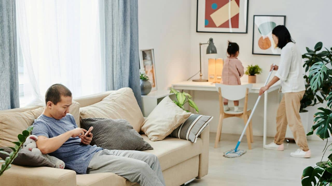 A man uses his phone on a sofa while a woman mops the floor nearby.