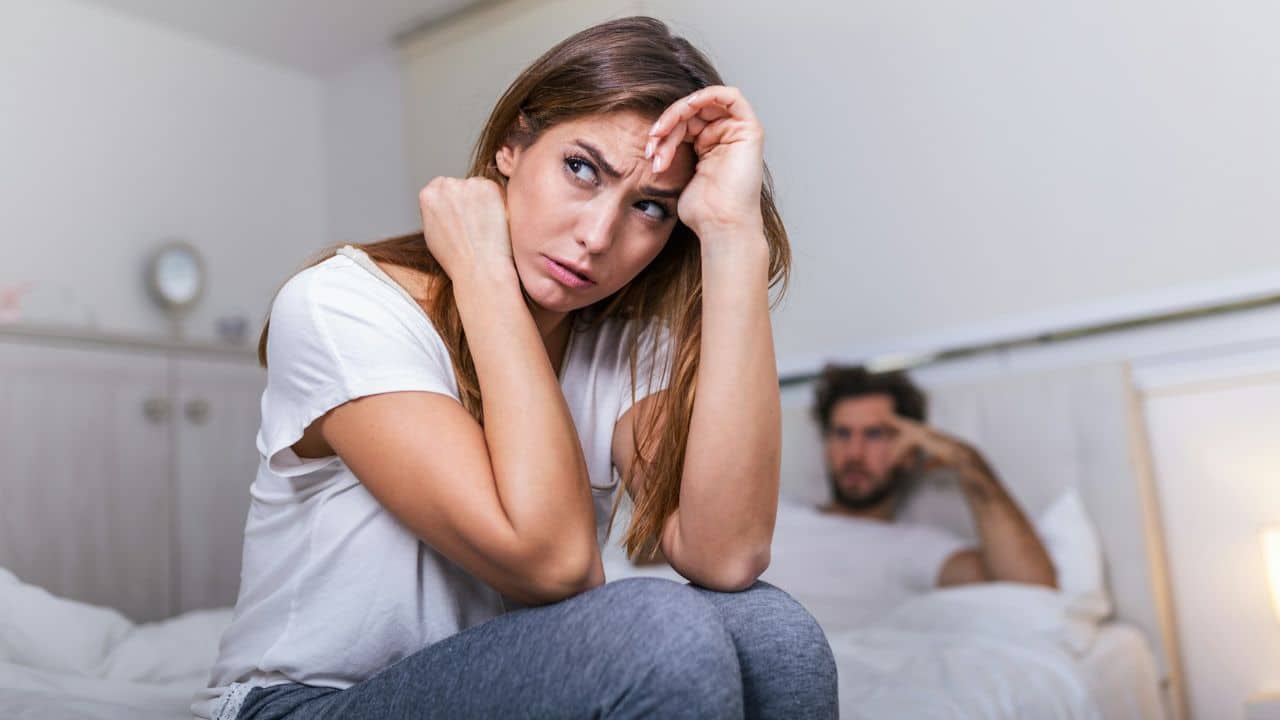 A woman sits on a bed with a worried expression while a man sits behind.