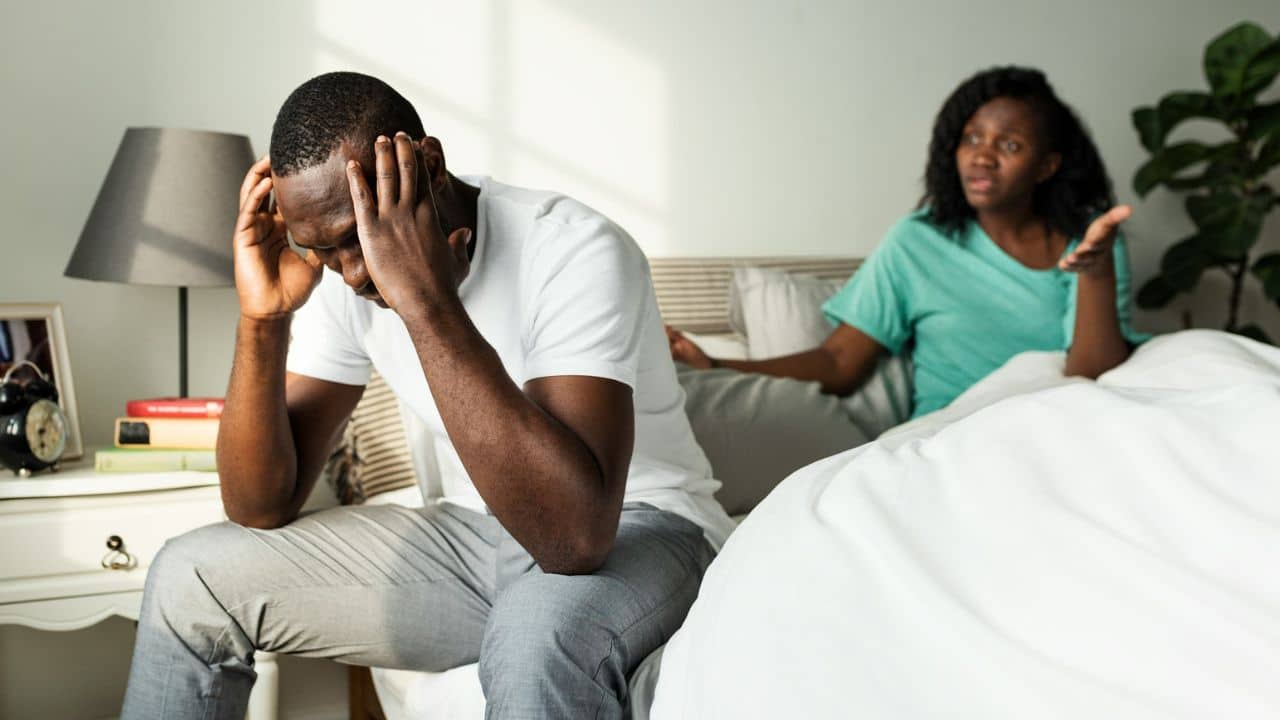 A man sits with his head in his hands while a woman gestures from the bed.